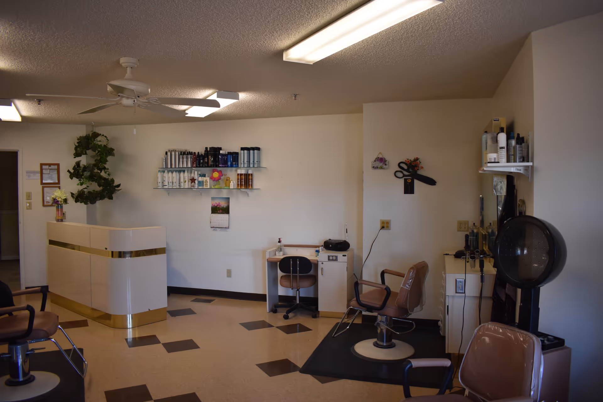 Interior view of a hair salon area within a senior living facility, featuring salon chairs, a hair dryer, shelves with hair care products, a reception desk, and a small workstation with a chair. The room has a ceiling fan and fluorescent lighting.