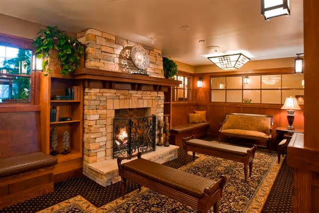 Cozy living room area with a stone fireplace, a fire burning inside, wooden benches with cushioned seats, a patterned rug, wooden paneling on the walls, built-in shelves with plants and decorative items, and warm lighting fixtures.