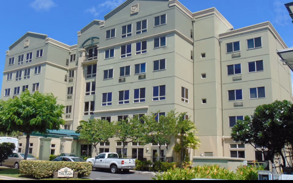 Front exterior of a multi-story pale green senior living building with parked cars and landscaping.