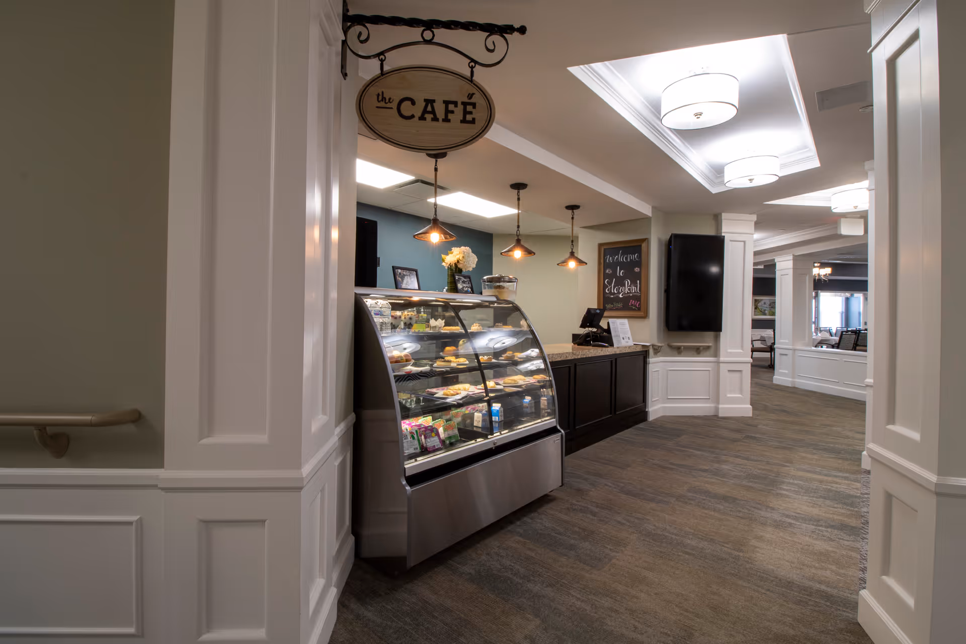 Interior view of a café area inside a senior living facility with a glass display case filled with pastries and snacks. The café sign hangs above the display case, and there is a counter with a cash register. The hallway is well-lit with ceiling lights and has carpeted flooring and white paneled walls.