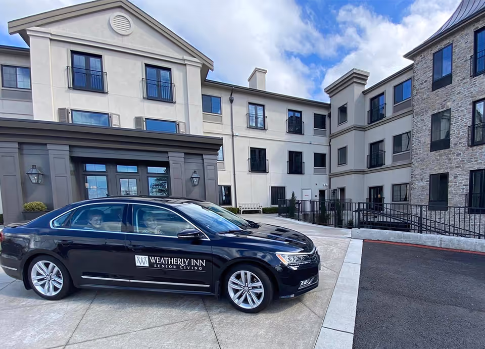 Exterior view of a senior living facility named Weatherly Inn Renton with a black car parked in front displaying the facility's logo. The building is three stories tall with multiple windows and a mix of stone and stucco exterior walls under a partly cloudy sky.