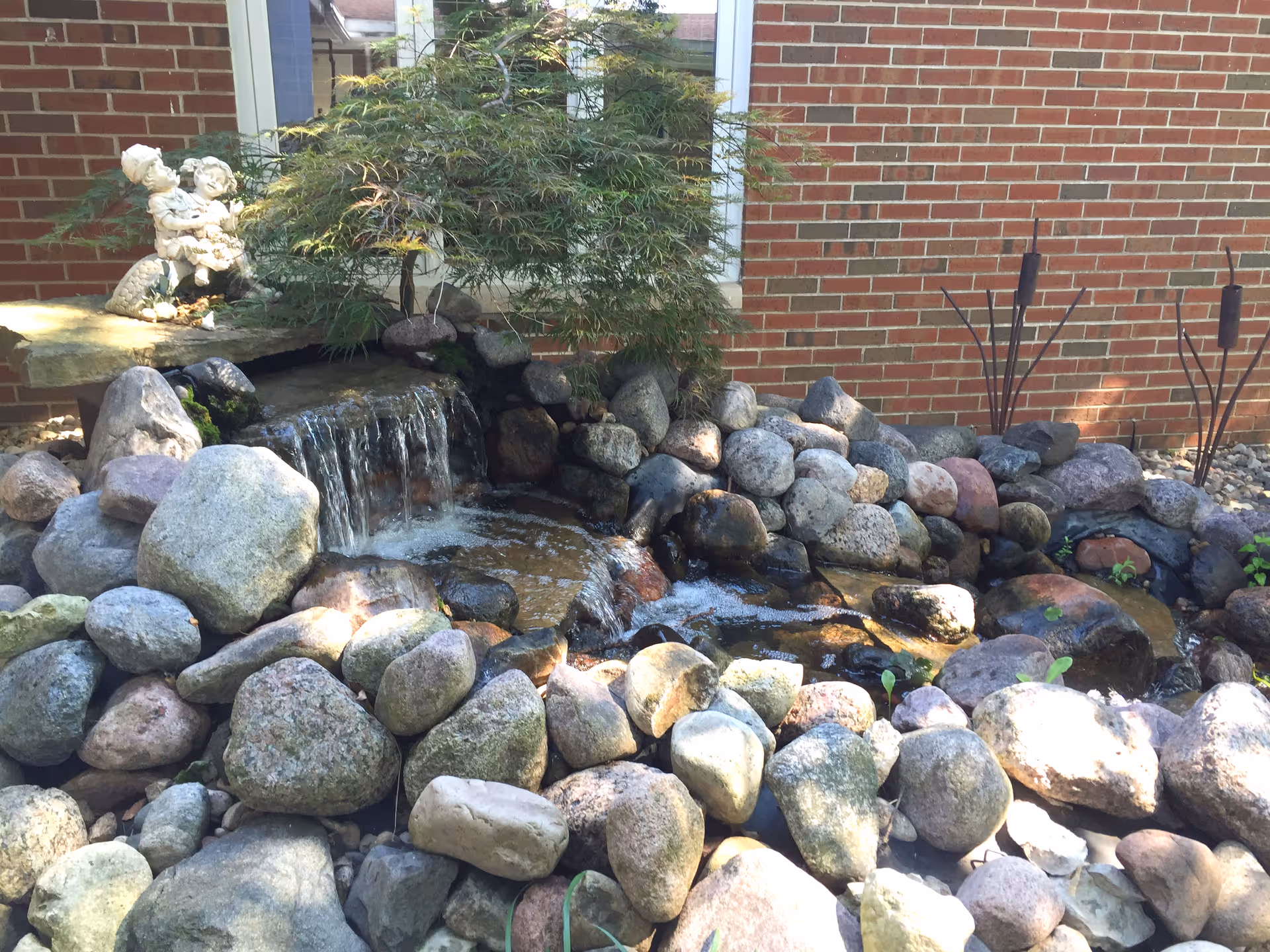 A small outdoor rock garden with a water feature including a small waterfall flowing over rocks. There is a green leafy plant above the waterfall and a brick wall in the background. A decorative statue of two children sitting together is placed on a flat stone near the waterfall.