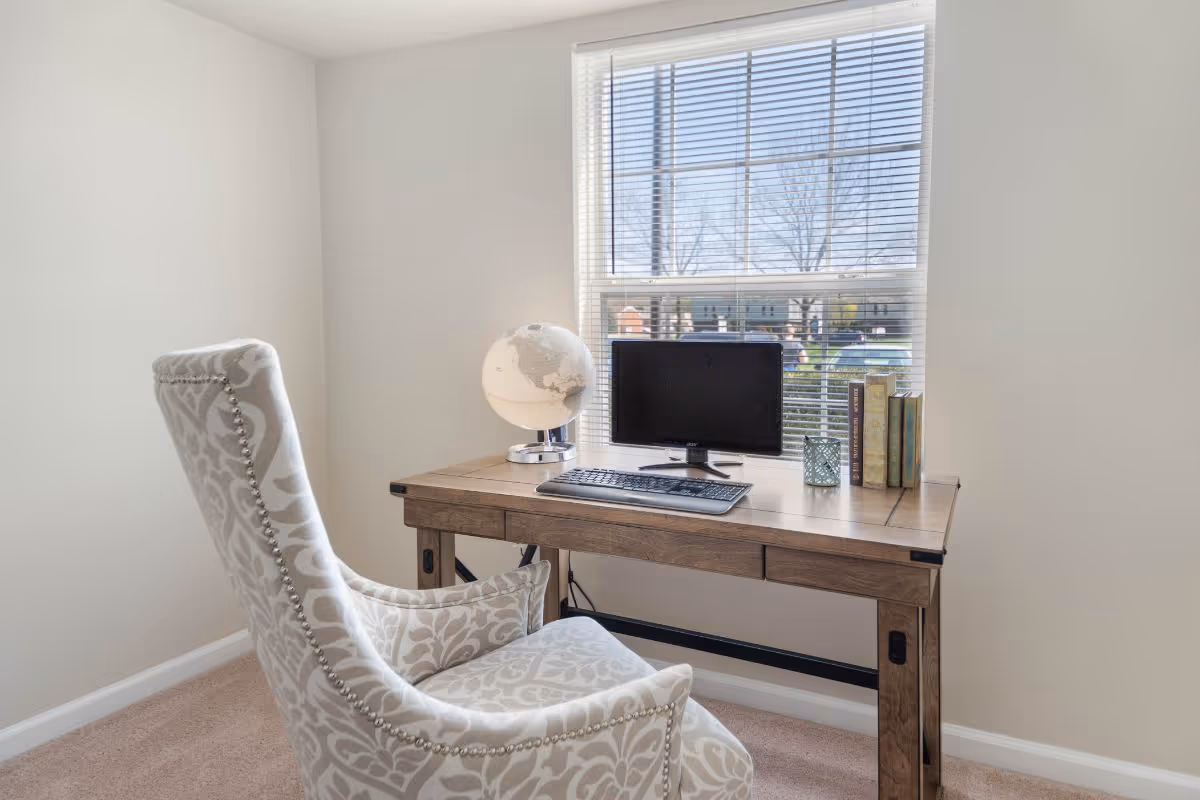 A small home-office corner with a wooden desk under a window, a monitor and keyboard, a globe and books, and a patterned upholstered armchair.