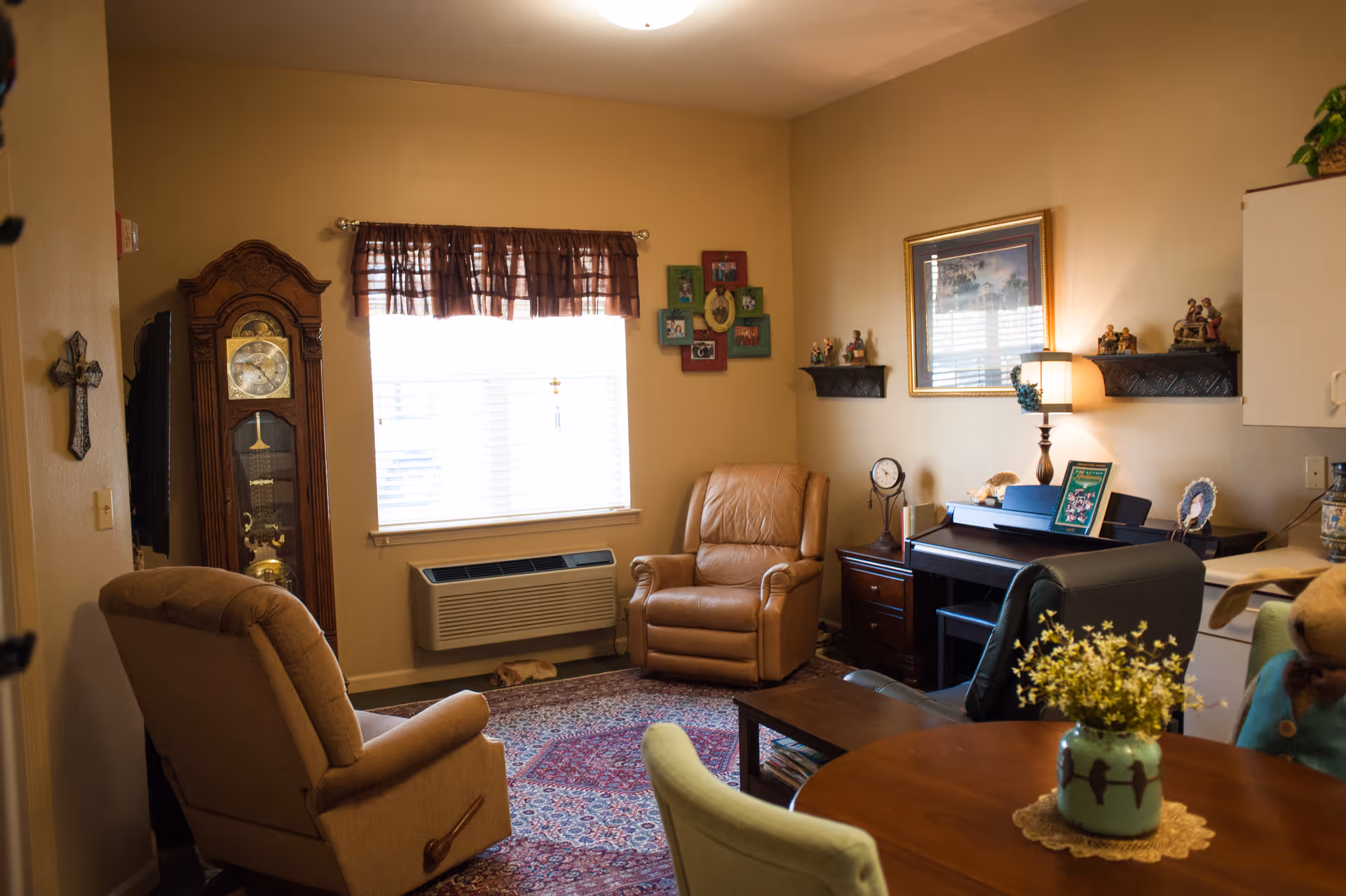 A cozy living room with beige walls and carpet. The room features a large window with brown curtains, a grandfather clock, two armchairs, a piano with a lamp and framed pictures on the wall above it, and a round wooden table with a vase of flowers in the foreground.