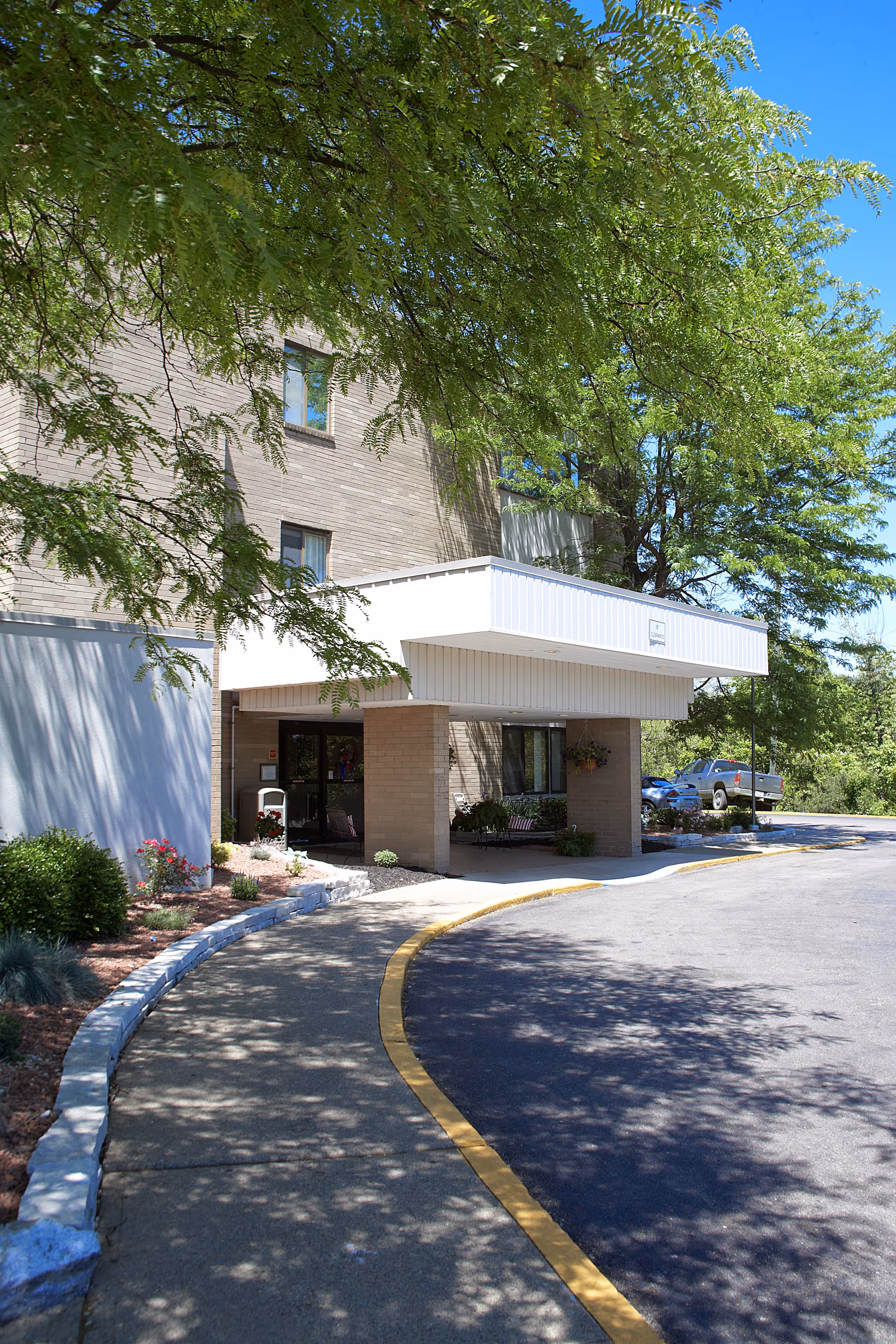Entrance to a multi-story brick building with a covered drop-off area. There is a curved sidewalk bordered by a low stone edge and landscaping with bushes and flowers. Trees provide shade over the area, and a few parked cars are visible in the background under a clear blue sky.