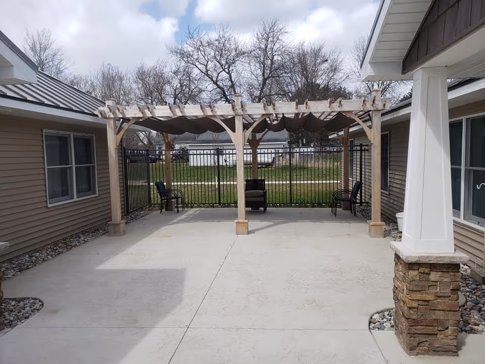 Outdoor patio area with a wooden pergola featuring a fabric canopy. There are three chairs placed under the pergola. The patio is surrounded by beige siding buildings and a black metal fence, with leafless trees and a cloudy sky in the background.