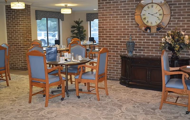 Dining area with round tables and wooden chairs with blue cushions, set with cups and napkins. The room has brick walls, a large decorative clock, a dark wooden sideboard with a vase and floral arrangement, and large windows with valances letting in natural light.
