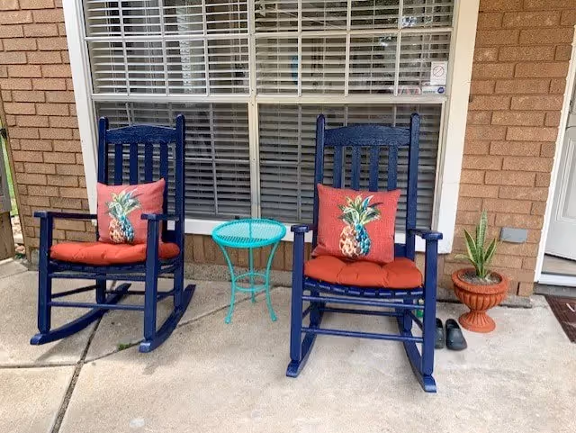 Two navy blue rocking chairs with red pineapple pillows flank a turquoise table on a porch.