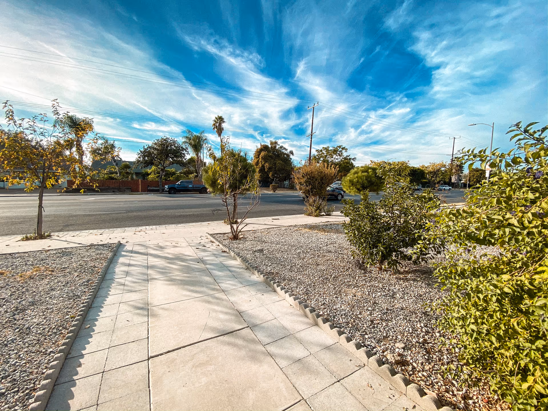 A paved walkway bordered by small rocks and various green shrubs and small trees leading to a street with cars and utility poles under a blue sky with wispy clouds.