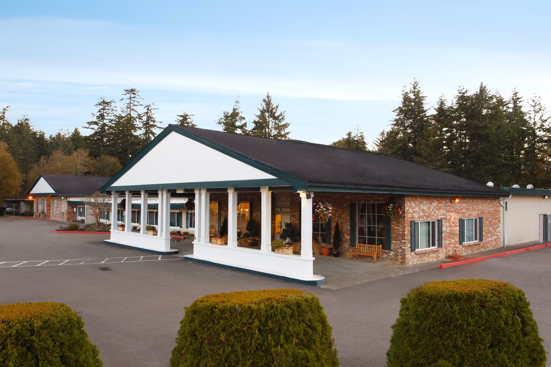 Exterior view of a single-story brick building with a covered entrance supported by white columns. The building is surrounded by a paved parking area and greenery, with tall trees in the background under a clear sky.