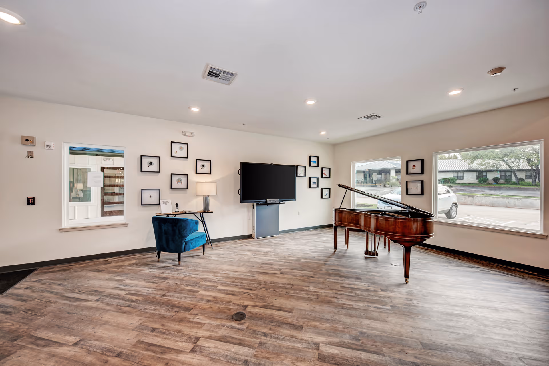 A spacious room with wooden flooring featuring a grand piano near large windows that let in natural light. There is a blue armchair next to a small table with a lamp and framed pictures on the walls. A large flat-screen TV is mounted on the wall.