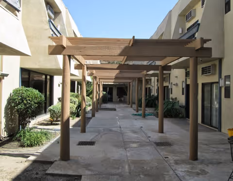 Outdoor courtyard area with a wooden pergola structure providing partial shade. The courtyard is surrounded by beige buildings with windows and some greenery including bushes and plants along the edges.