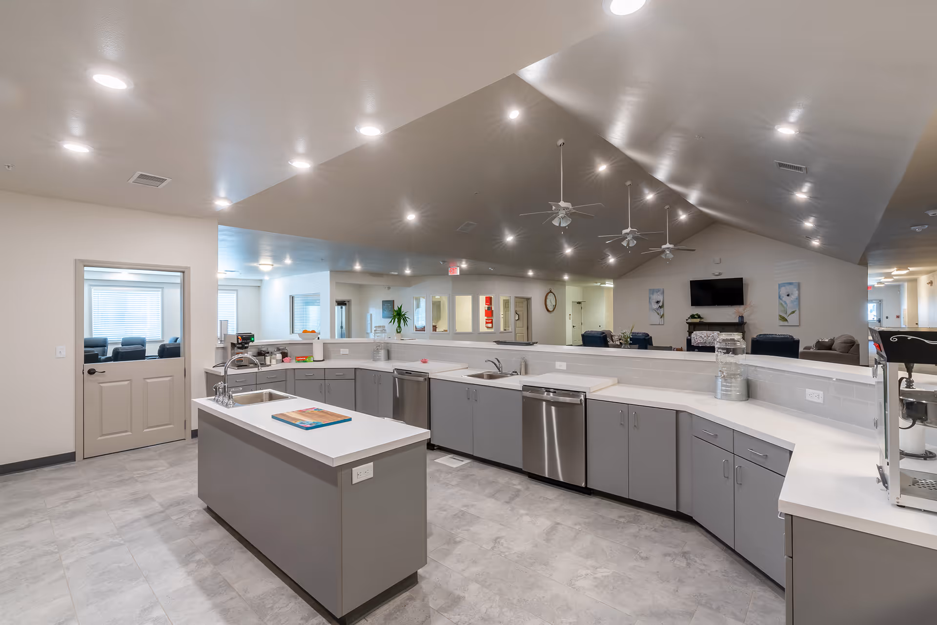 A spacious, modern kitchen area with gray cabinets and white countertops. The kitchen features two sinks, a dishwasher, and an island with a cutting board on top. The ceiling is vaulted with recessed lighting and ceiling fans. In the background, there is a living area with chairs, a TV mounted on the wall, and decorative artwork.