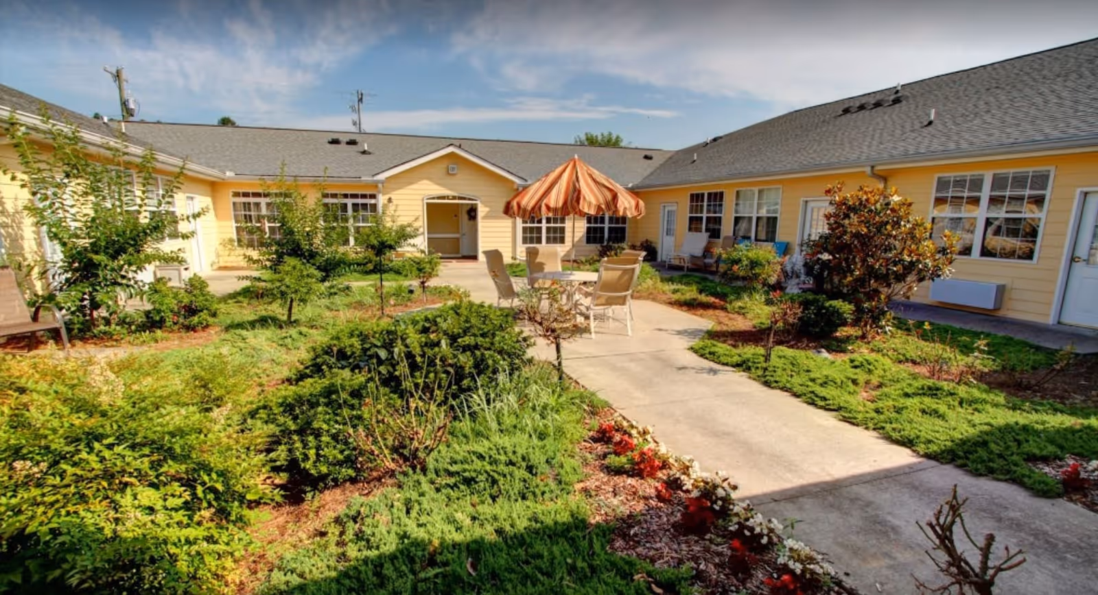 Outdoor courtyard area at Charter Senior Living of Hermitage featuring a paved walkway, green shrubs, small trees, and a round table with chairs under a striped umbrella. The courtyard is surrounded by a single-story yellow building with multiple windows and doors.