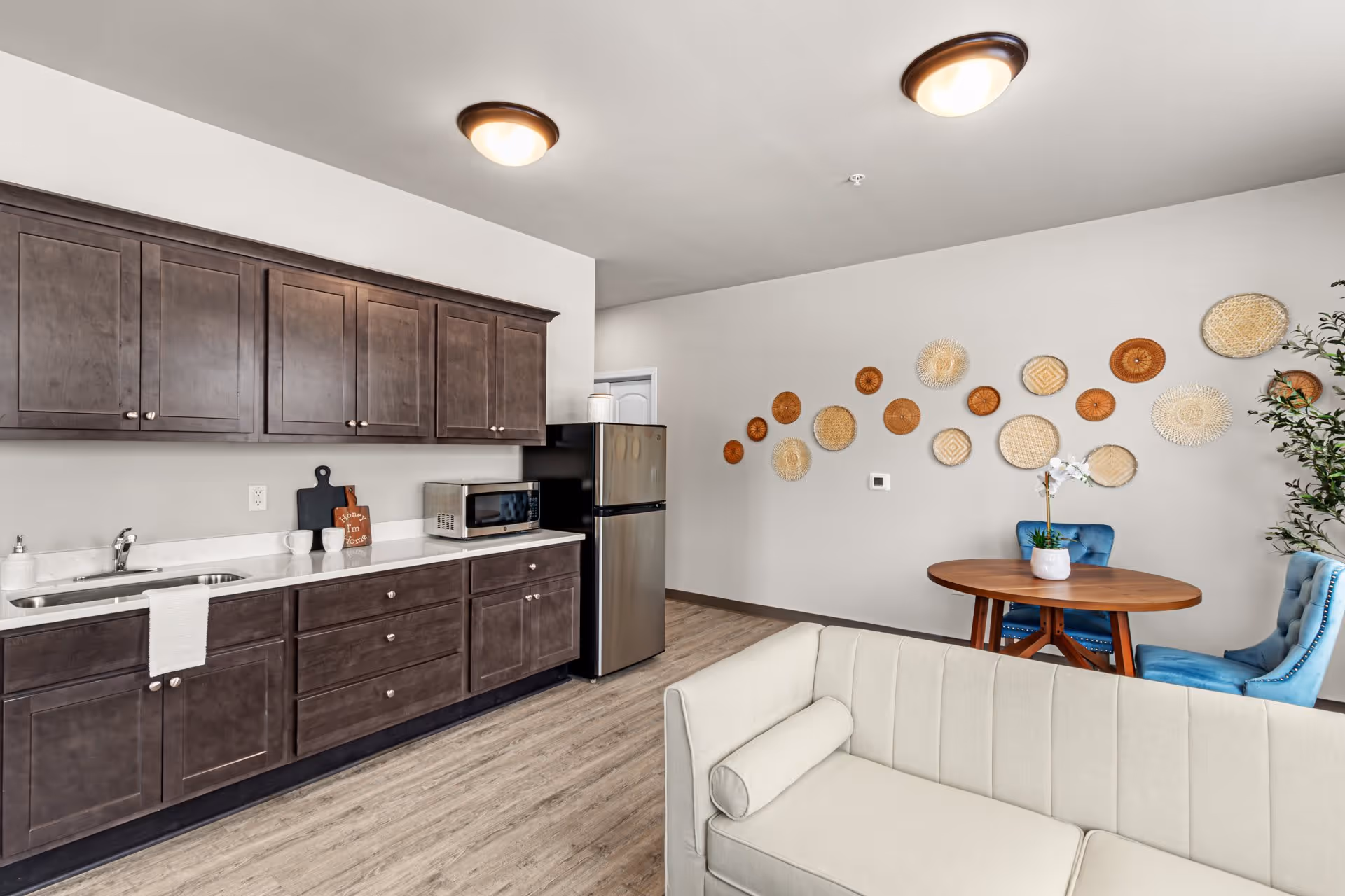 Interior view of a living space featuring a kitchenette with dark wood cabinets, a stainless steel refrigerator, a microwave, and a sink with a towel hanging on the cabinet. Adjacent to the kitchenette is a small round wooden table with two blue upholstered chairs and a white orchid plant on top. The wall behind the table is decorated with various woven basket wall hangings. In the foreground, there is a light beige sofa with a cylindrical pillow. The floor is wood laminate and the ceiling has two round light fixtures.