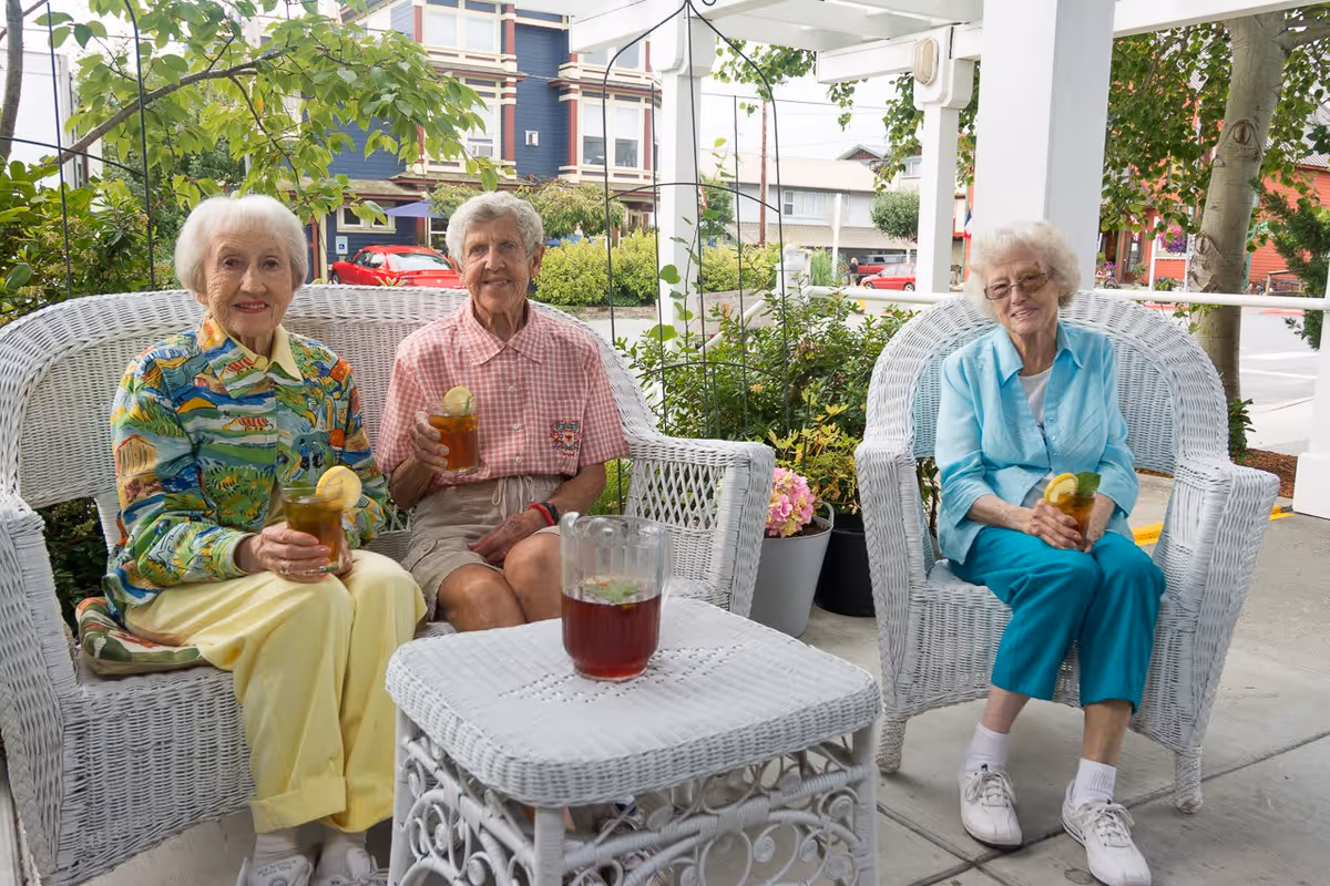 Three elderly women sitting on white wicker chairs on a covered porch, each holding a glass of iced tea with lemon. There is a white wicker table in front of them with a pitcher of iced tea. Green plants and a street with buildings and cars are visible in the background.