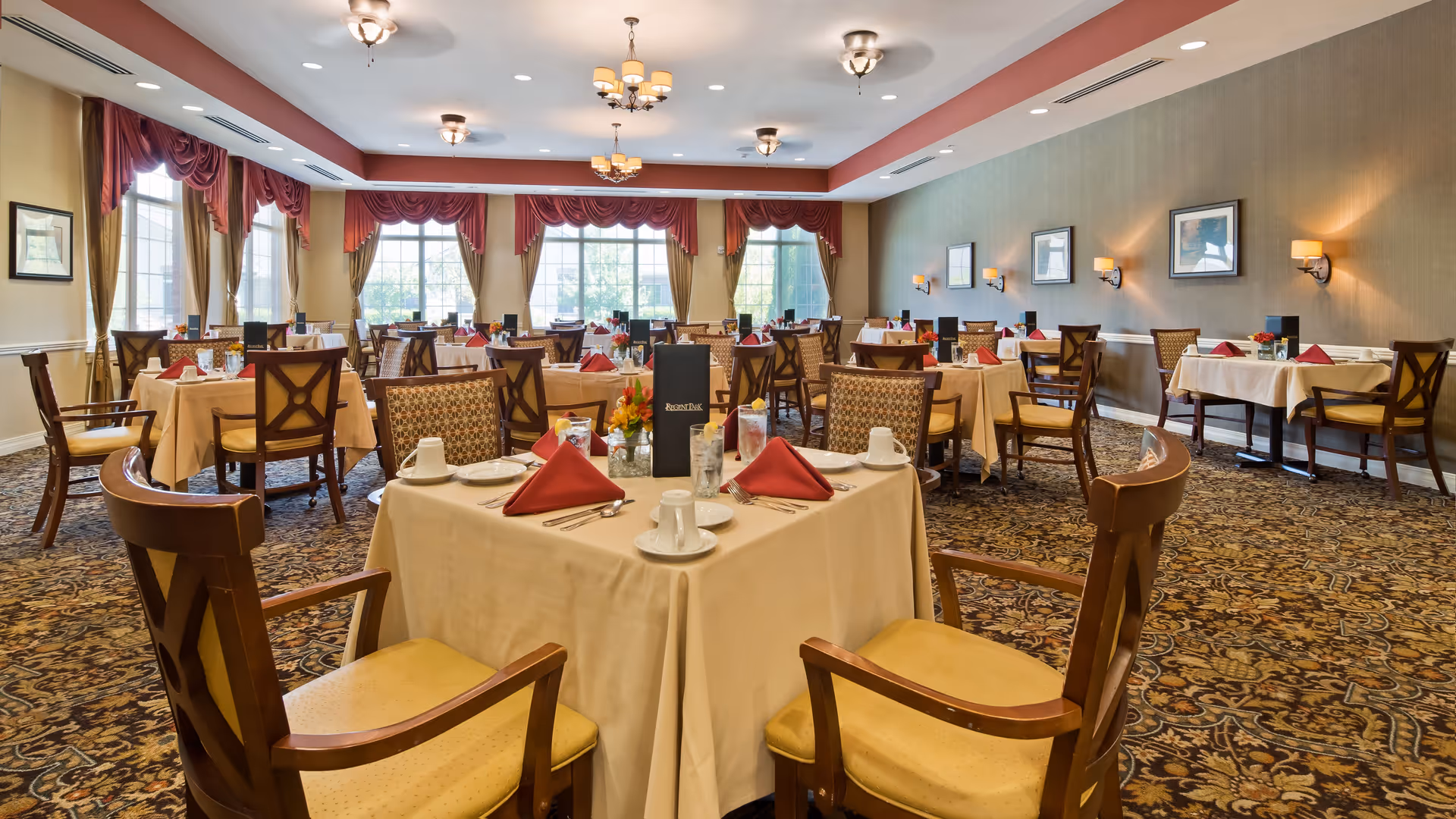 A spacious dining room with multiple tables covered in beige tablecloths, each set with white cups, plates, silverware, and red folded napkins. The room features large windows with red curtains, patterned carpet, and warm lighting from ceiling fixtures and wall sconces.