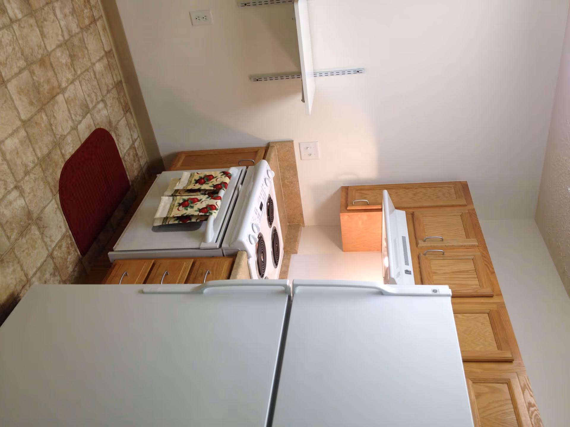 Small kitchen with a white refrigerator and stove, wooden cabinets, tiled floor and a red rug.