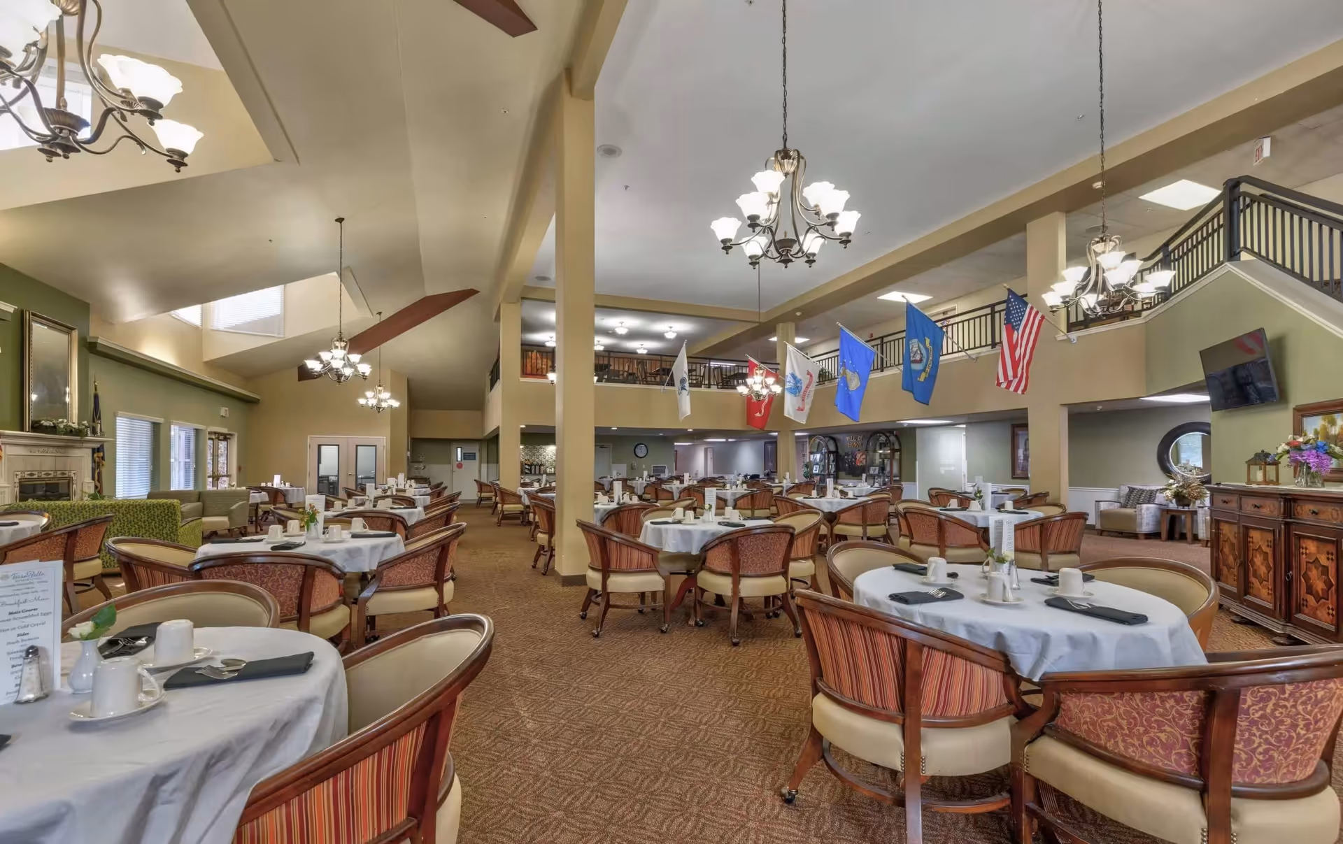 Spacious dining room with multiple round tables covered in white tablecloths, each set with cups, napkins, and utensils. Comfortable upholstered chairs surround the tables. The room features high ceilings with chandeliers, flags hanging from the upper level railing, and a cozy seating area with a fireplace and large windows on one side.