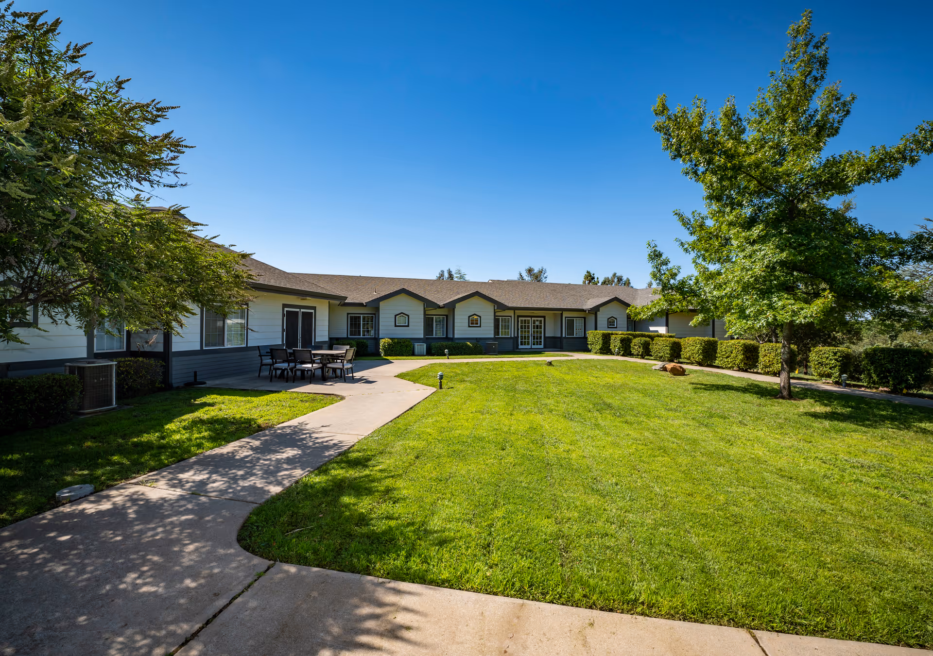 A sunny outdoor courtyard area at Country Crest Post-Acute featuring a well-maintained green lawn, a concrete walkway, several trees, and a single-story building with multiple windows and doors. There is a patio area with a table and chairs on the left side.