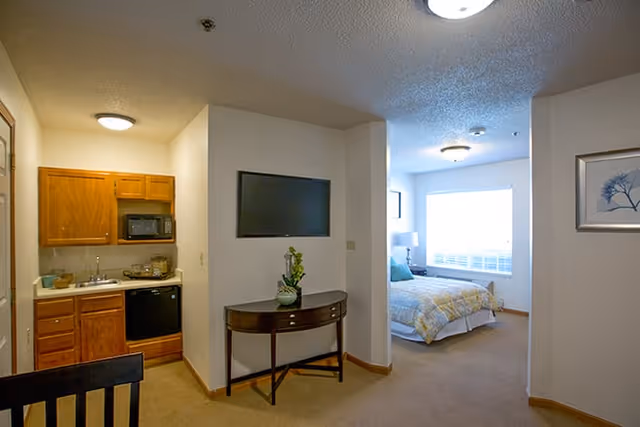 Interior view of a senior living facility apartment at Brookdale Columbia showing a small kitchen area with wooden cabinets, a microwave, and a mini refrigerator on the left. In the center, there is a wall-mounted flat-screen TV above a small wooden console table with a plant. To the right, a bedroom area is visible with a bed, a bedside lamp, and a large window letting in natural light.