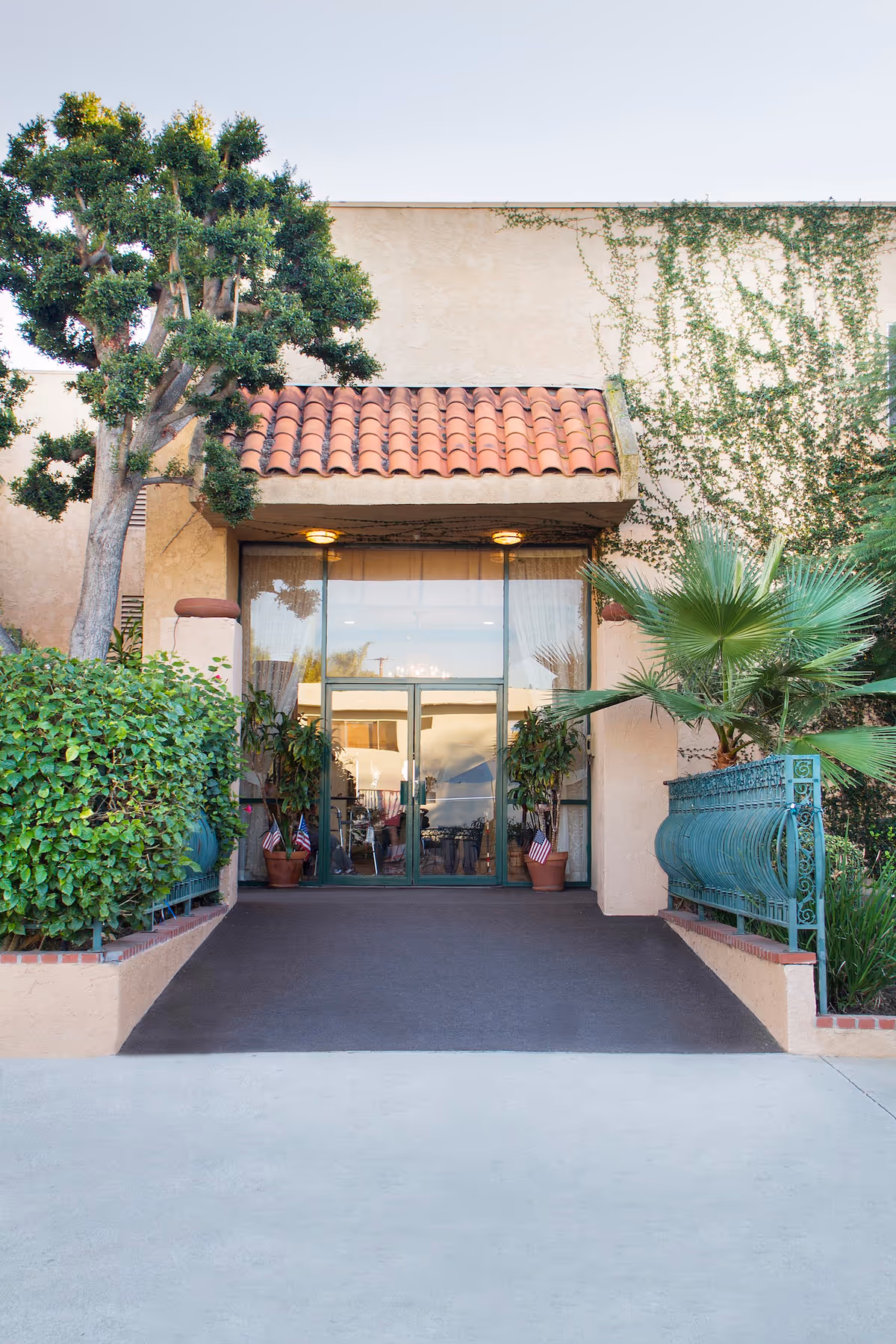 Entrance to a building with a tiled roof overhang, glass double doors, and potted plants on either side. The building exterior is beige with some greenery and a tree on the left side.
