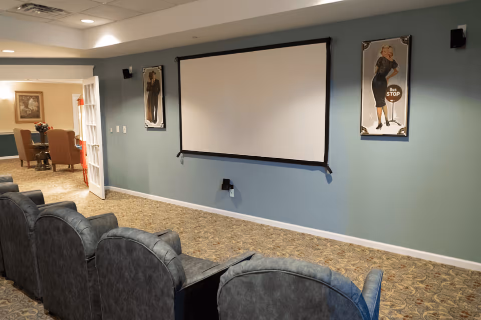 Small screening room with upholstered theater chairs facing a wall-mounted projection screen and framed art on the walls.