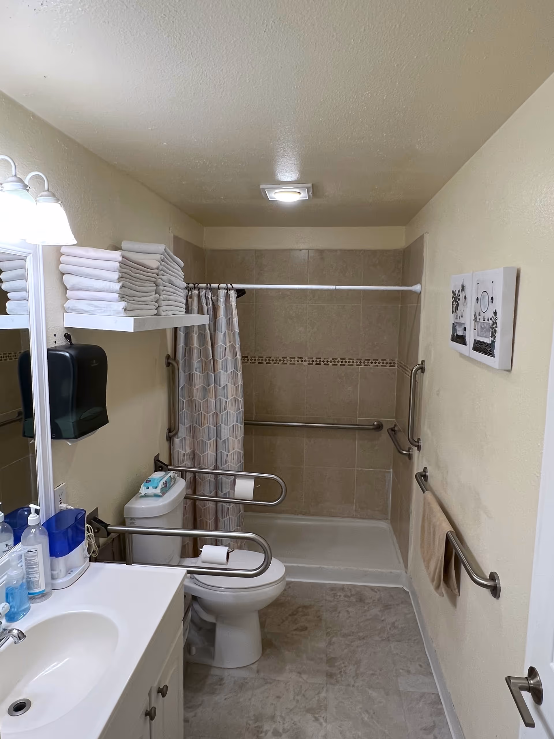 A clean and accessible bathroom with a white sink and vanity on the left, a toilet with safety grab bars around it, and a walk-in shower with a curtain and tiled walls. There are multiple grab bars installed on the walls for support, a shelf with neatly folded white towels above the toilet, and two framed pictures on the right wall. The floor has a light-colored tile, and the bathroom is well-lit with ceiling and wall lights.