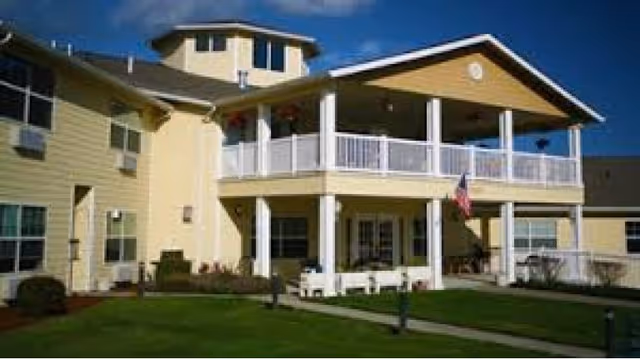 Exterior view of a two-story senior living facility building with a covered balcony on the second floor, white railings, and a well-maintained lawn in front. The building is painted yellow with white trim and has several windows. An American flag is displayed near the entrance.