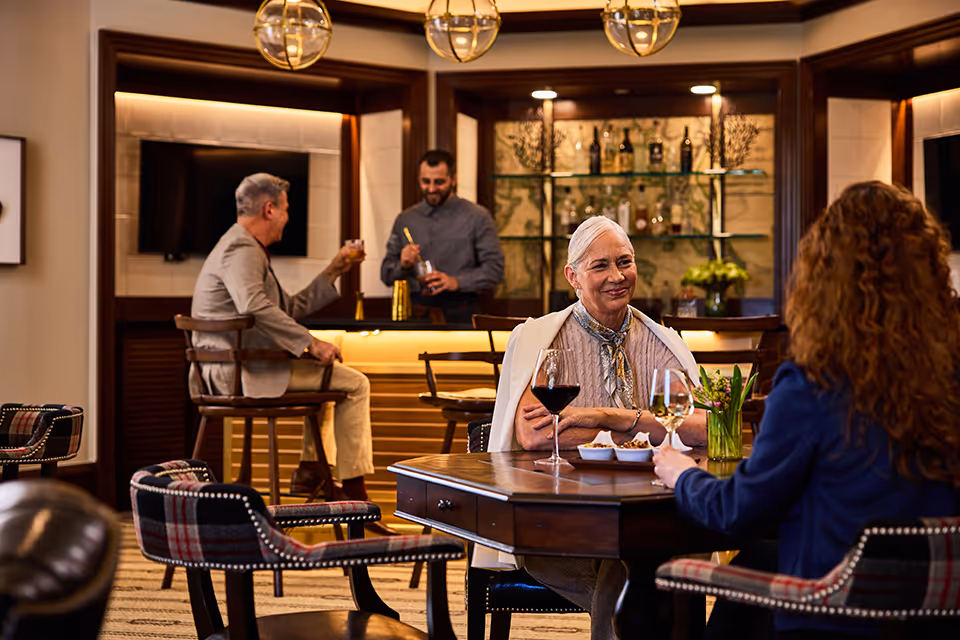 Two women sitting at a wooden table in a cozy lounge area, enjoying drinks and snacks. In the background, a man is sitting on a bar stool holding a drink while another man behind the bar is preparing a cocktail. The room features warm lighting, wooden furniture, and a decorative bar with bottles and plants.