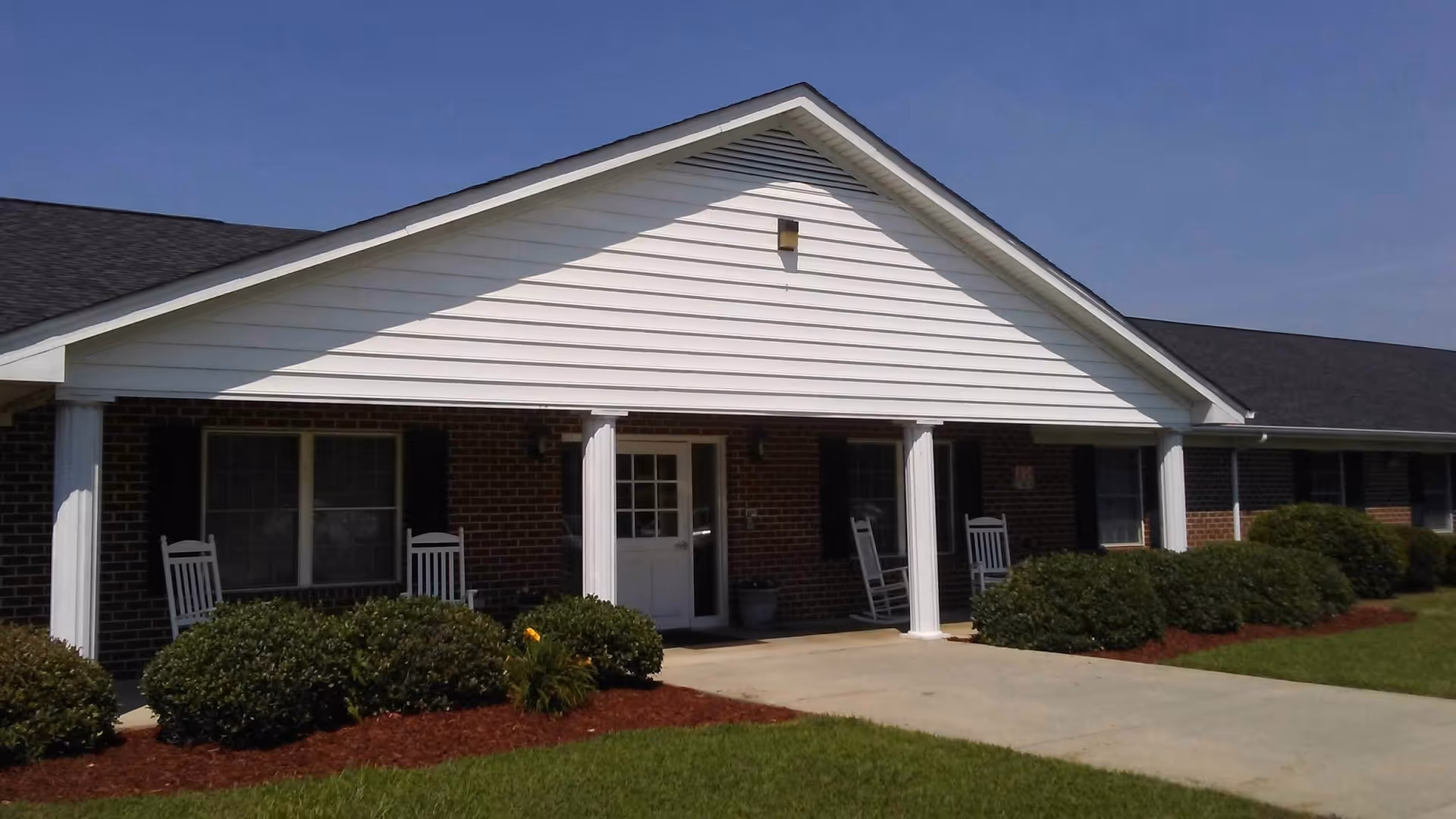 Front exterior view of Hampton Manor, a single-story brick building with white columns and a covered porch featuring rocking chairs. The entrance is centered with windows on either side, surrounded by neatly trimmed bushes and a well-maintained lawn under a clear blue sky.