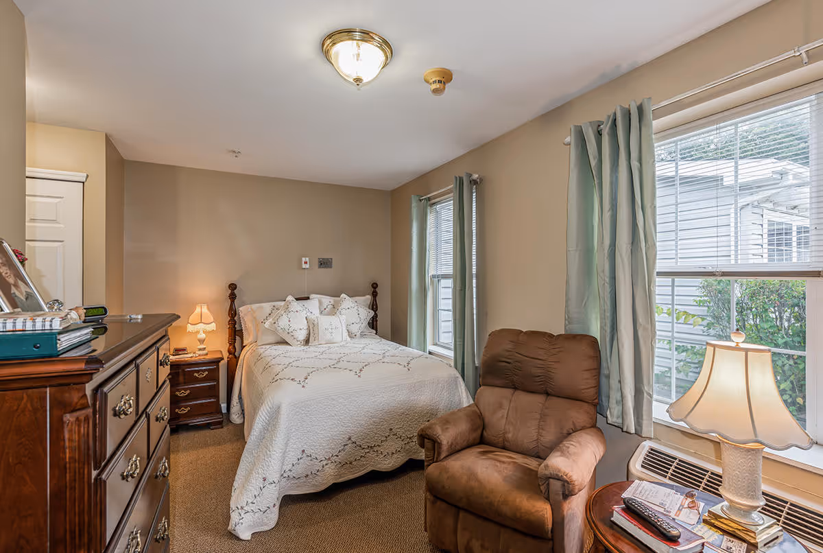 A cozy bedroom with a double bed covered in a white quilt and decorative pillows. There is a wooden nightstand with a lamp beside the bed, a wooden dresser with items on top, a comfortable brown recliner chair near two windows with light green curtains, and a small round table with a lamp and some books and papers.