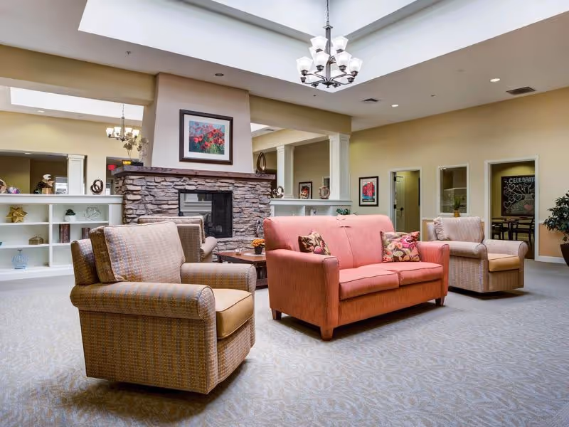 A cozy living room area in a senior living facility featuring a stone fireplace with a framed floral painting above it. The room has a pink sofa with floral cushions, two beige armchairs, a wooden coffee table, built-in white shelves with decorative items, and a chandelier hanging from the ceiling. The walls are painted a warm beige color and there are doorways leading to other rooms.