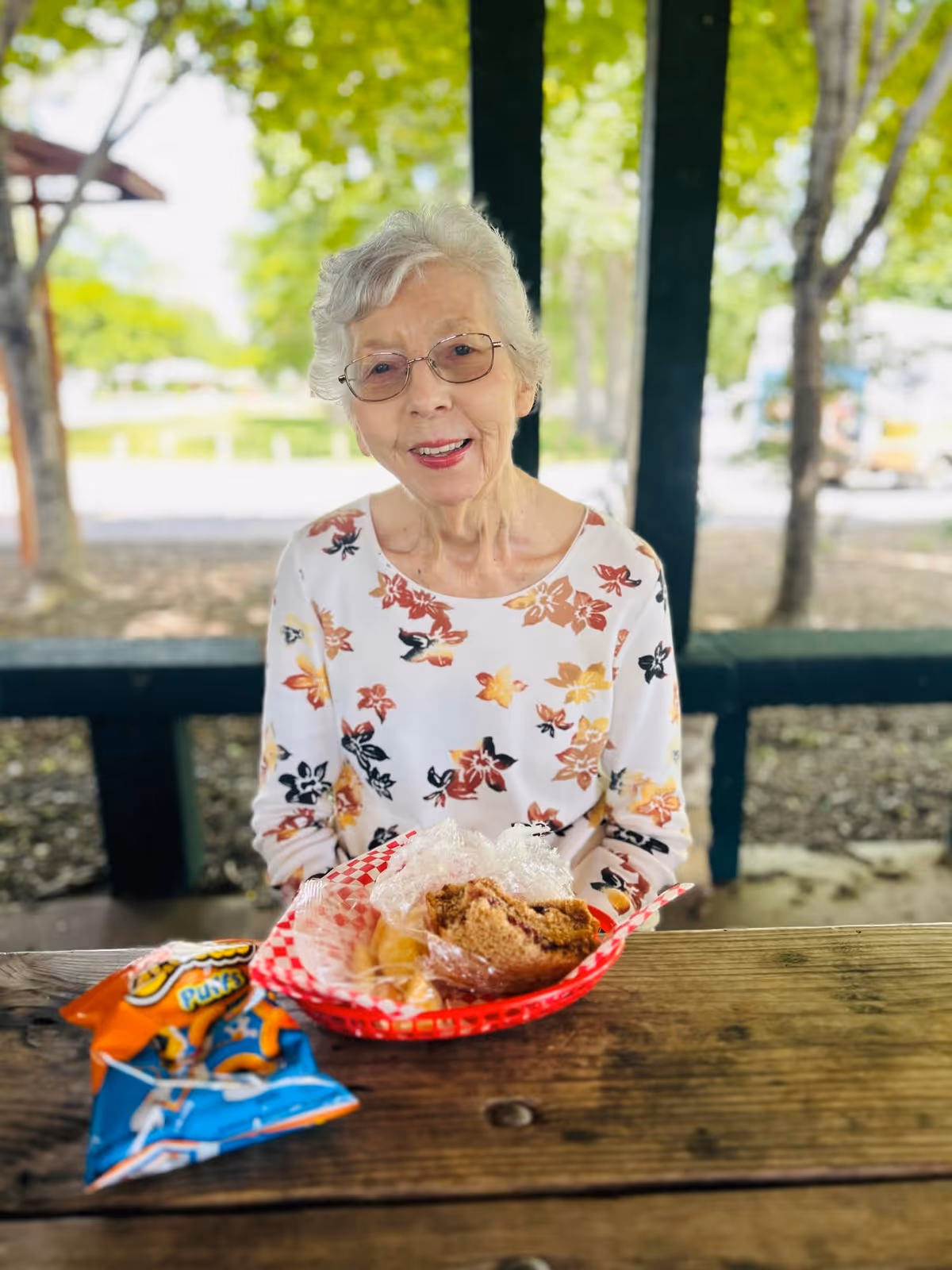 An elderly woman with short gray hair and glasses is sitting at a wooden picnic table outdoors. She is wearing a white shirt with a floral pattern in shades of orange, yellow, and black. In front of her on the table is a red basket with a sandwich wrapped in plastic and some chips, along with two small bags of snacks. The background shows trees and a park-like setting with sunlight filtering through the leaves.