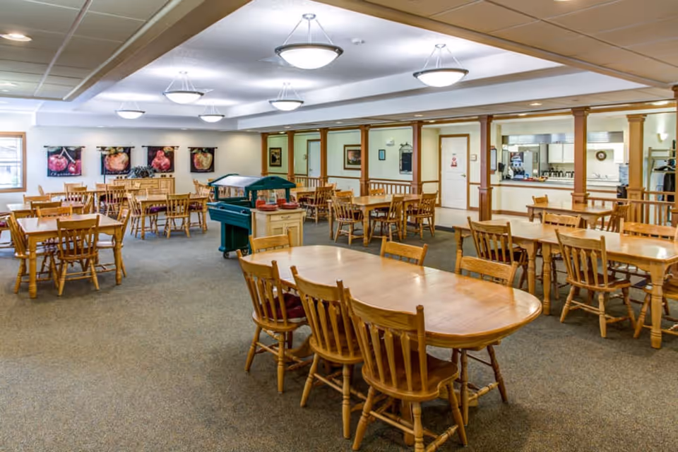 A spacious dining room with multiple wooden tables and chairs arranged neatly. The room has carpeted flooring, ceiling lights, and artwork on the walls. There is a serving area with a window to the kitchen in the background.