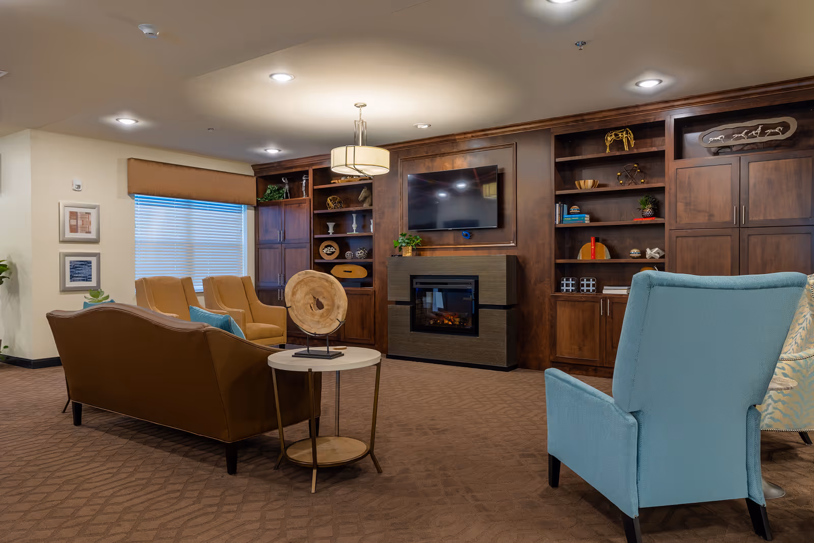 A cozy living room area with a brown leather sofa, two mustard yellow armchairs, and a light blue armchair. The room features a built-in wooden shelving unit with decorative items, a flat-screen TV mounted above a modern electric fireplace, and a round side table with a decorative wooden piece. The carpet is brown with a subtle pattern, and the ceiling has recessed lighting and a hanging light fixture.