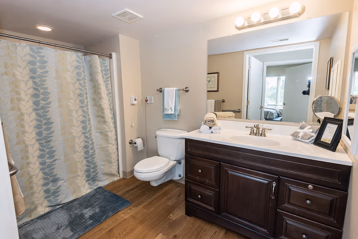 A clean and well-lit bathroom featuring a shower with a patterned curtain, a toilet, a dark wood vanity with a white countertop and sink, a large mirror above the sink with lights, and neatly folded towels on the countertop. The bathroom has wood-style flooring and light-colored walls.