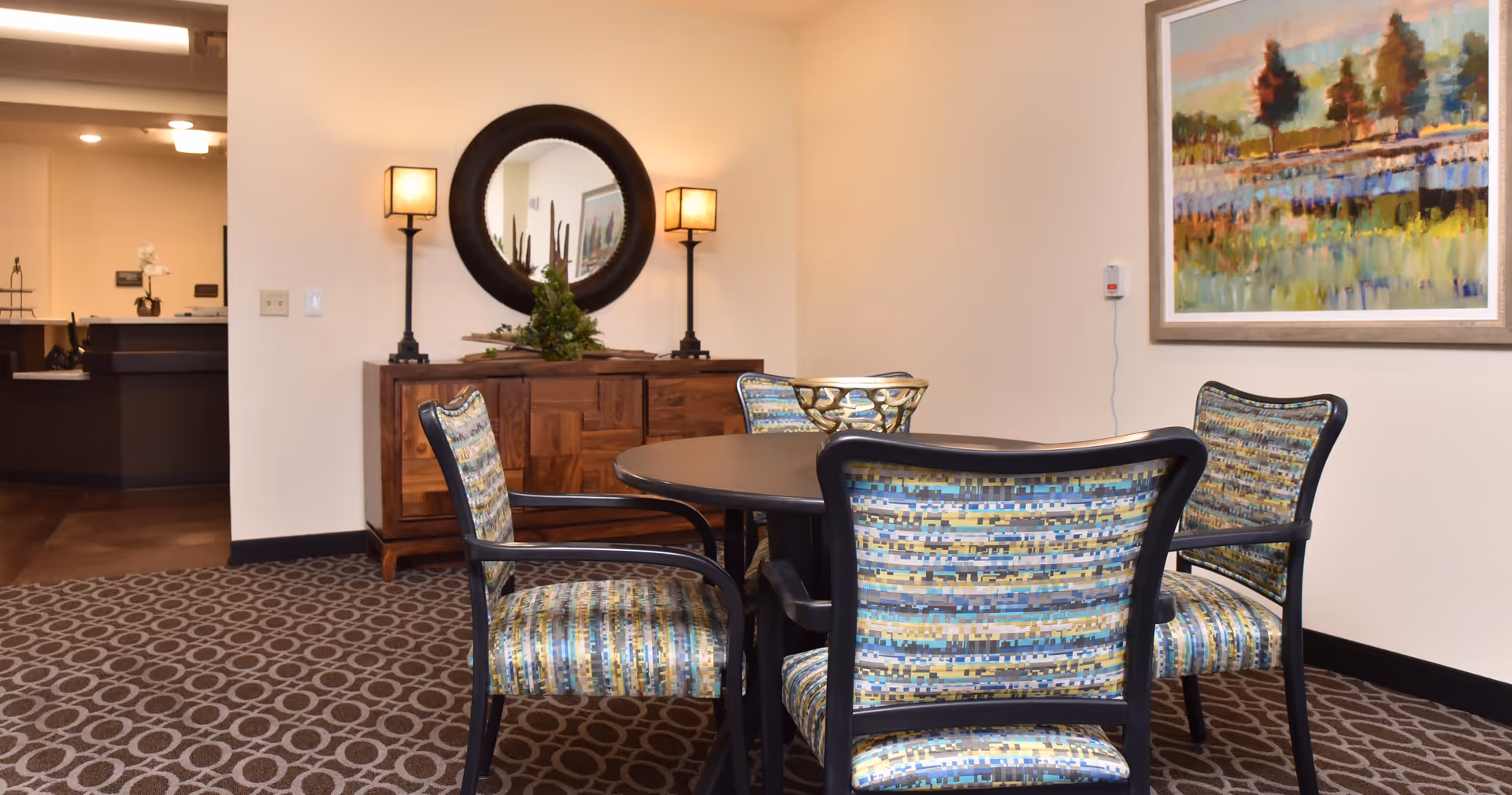 Interior seating area with a round table and four patterned chairs, a sideboard with a round mirror and lamps, and framed artwork on the wall.
