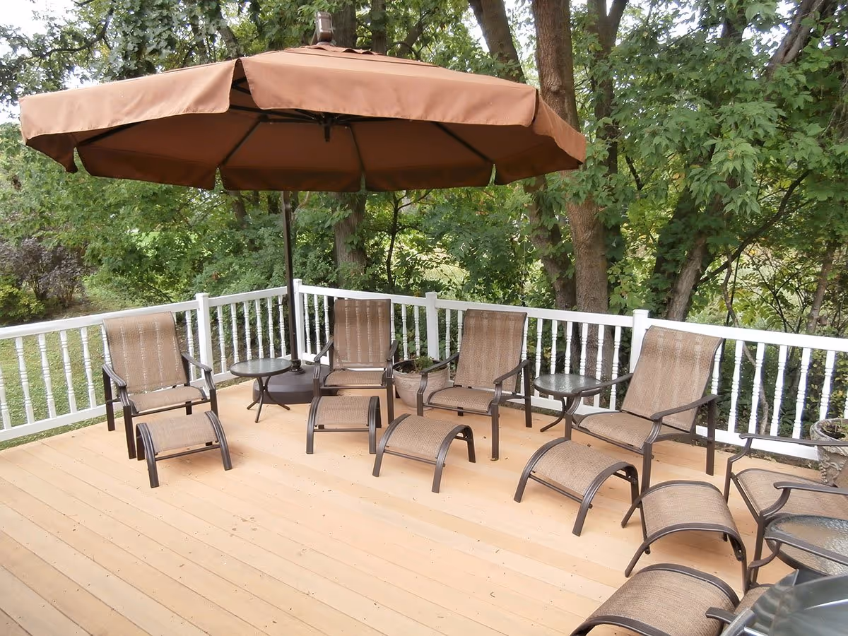 Wooden deck patio with brown lounge chairs, small tables, and a large brown umbrella overlooking trees.