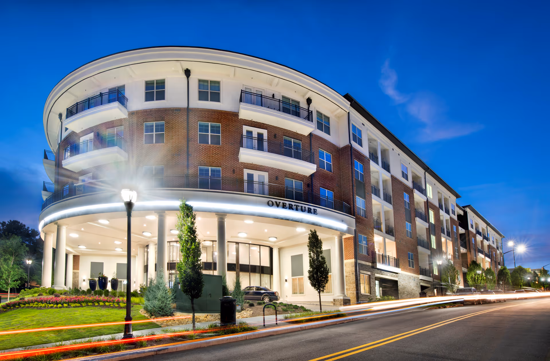 Evening exterior view of the curved multi-story brick building 'Overture' with a lit entrance, balconies, and landscaped frontage.
