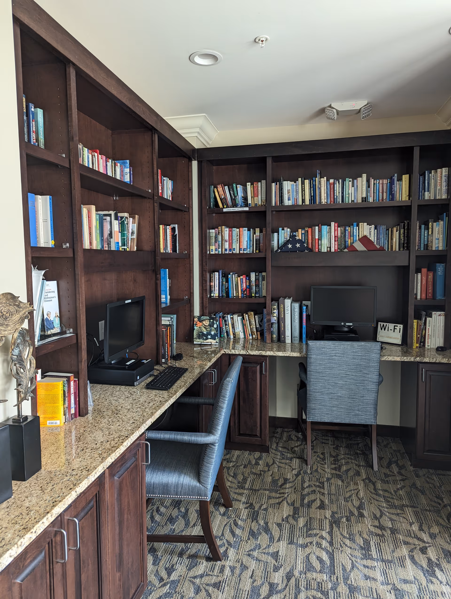 A cozy library and computer area with dark wooden bookshelves filled with books, two computer monitors on a granite countertop, and two gray upholstered chairs. The carpet has a patterned design, and there is a small sign indicating Wi-Fi availability.