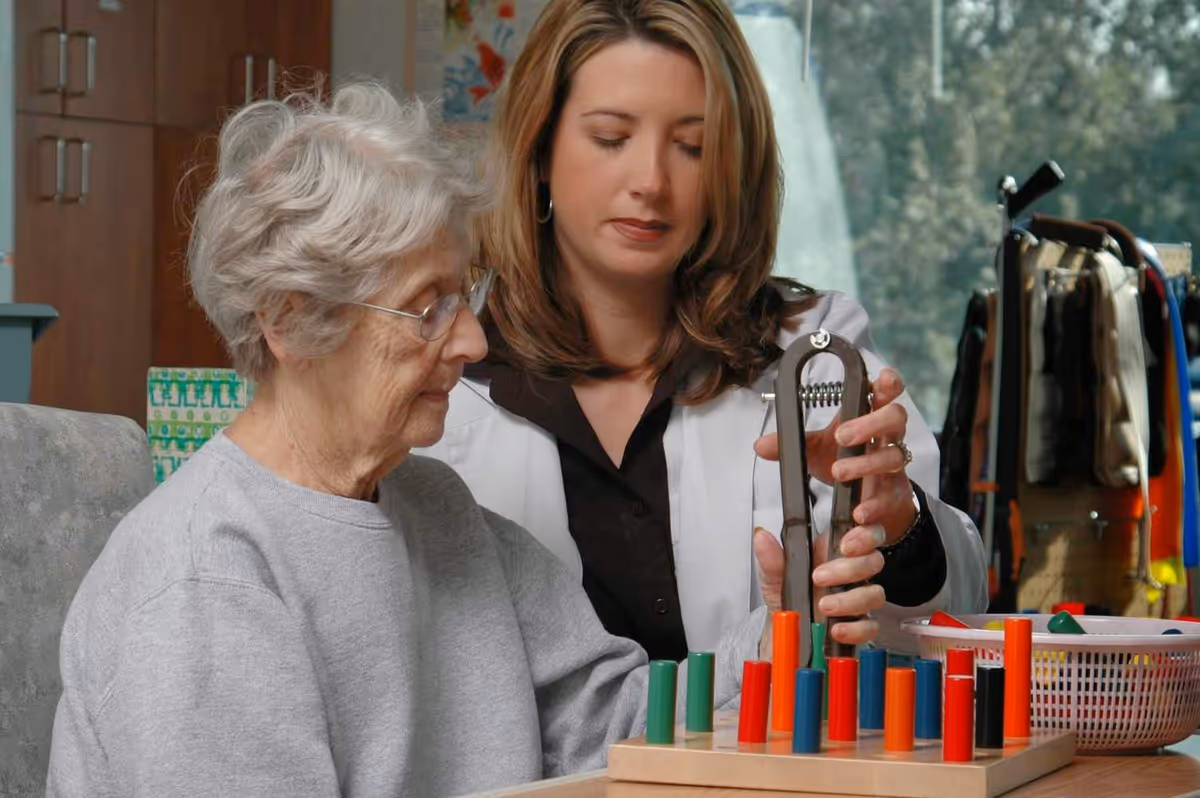 An elderly woman and a younger woman, possibly a therapist or caregiver, are engaged in a therapeutic activity using a pegboard with colorful pegs. The younger woman is assisting the elderly woman with a hand exercise device in a well-lit indoor setting.