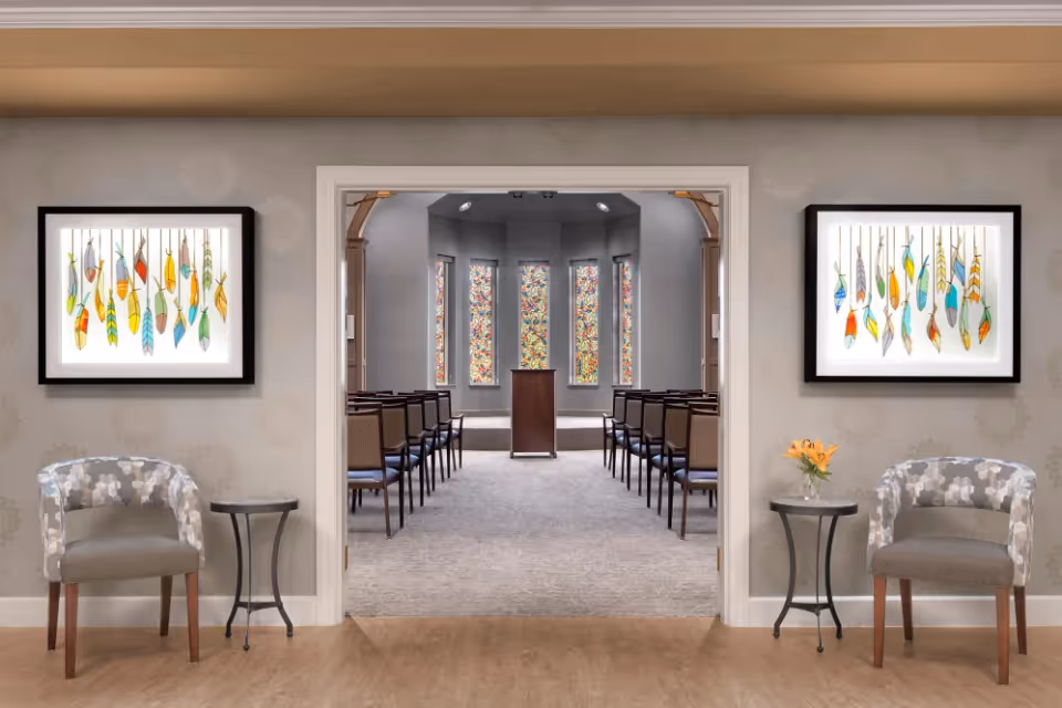 Entryway with two chairs and small tables framing a doorway that leads into a chapel-style room with rows of chairs, a podium, and stained-glass windows.