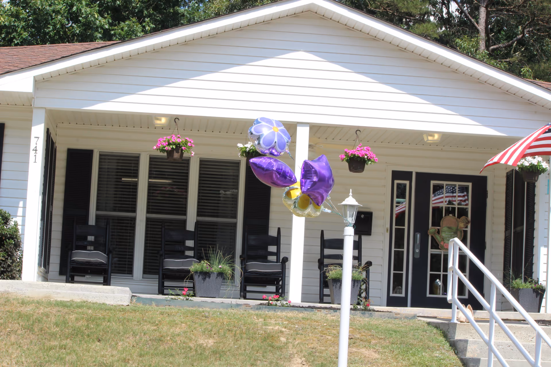 Front exterior view of a white single-story building with a covered porch featuring black rocking chairs, hanging flower baskets, potted plants, and purple and yellow balloons tied to a white lamp post. The entrance has double glass doors with black frames and an American flag reflected in the glass. A red and white striped umbrella is visible on the right side.