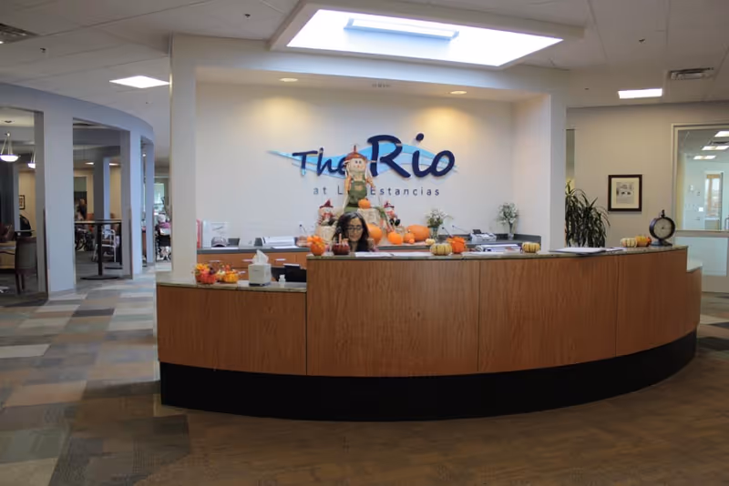 A curved reception desk in a senior living facility with a receptionist seated behind it, autumn pumpkin decorations, and a 'The Rio' sign on the wall.