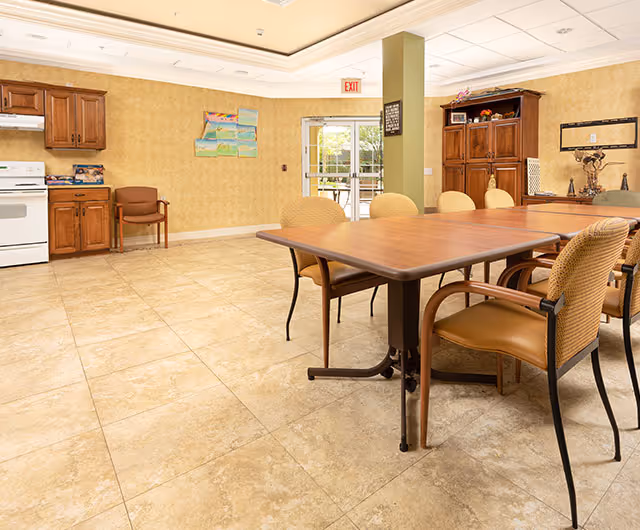 A spacious room with a large wooden table surrounded by six chairs with woven backs and cushioned seats. The room has beige tiled flooring and light yellow walls with a subtle pattern. On the left side, there is a small kitchen area with wooden cabinets, a white stove, and a chair. At the back, there is a glass door leading outside, and a wooden cabinet with decorative items on top is visible on the right side. The ceiling has recessed lighting and a tray design.