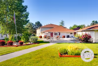 Outdoor view of a senior living facility with a well-maintained lawn, paved walkways, an American flag on a flagpole, patio tables and chairs, and buildings with red roofs surrounded by trees and shrubs under a clear blue sky.