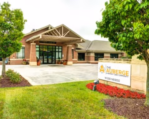 Front exterior of The Auberge at Oak Village assisted living facility showing the main entrance, driveway, landscaped lawn, and a sign.