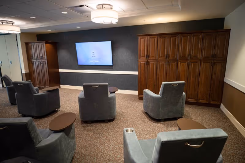 A lounge-style room with upholstered armchairs arranged facing a wall-mounted TV and wooden cabinetry.