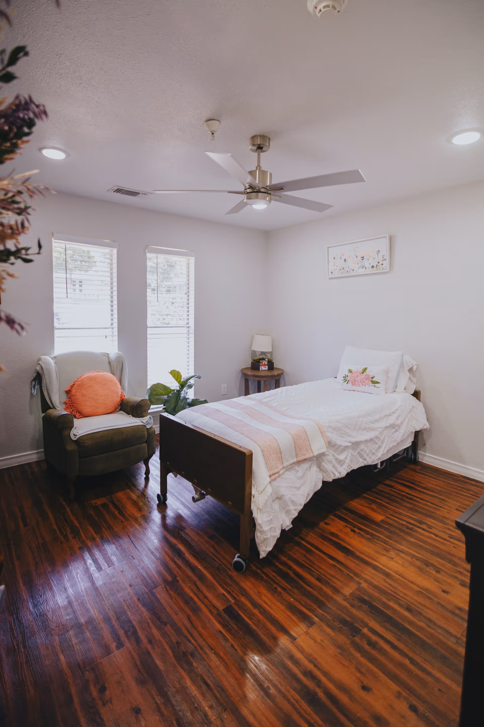 A cozy bedroom with a single bed covered in white bedding and a pink striped blanket. There is a green armchair with an orange round pillow near two windows with blinds. A small round wooden side table with a lamp and a small plant is next to the bed. The room has wooden flooring, a ceiling fan, and a framed floral artwork on the wall.
